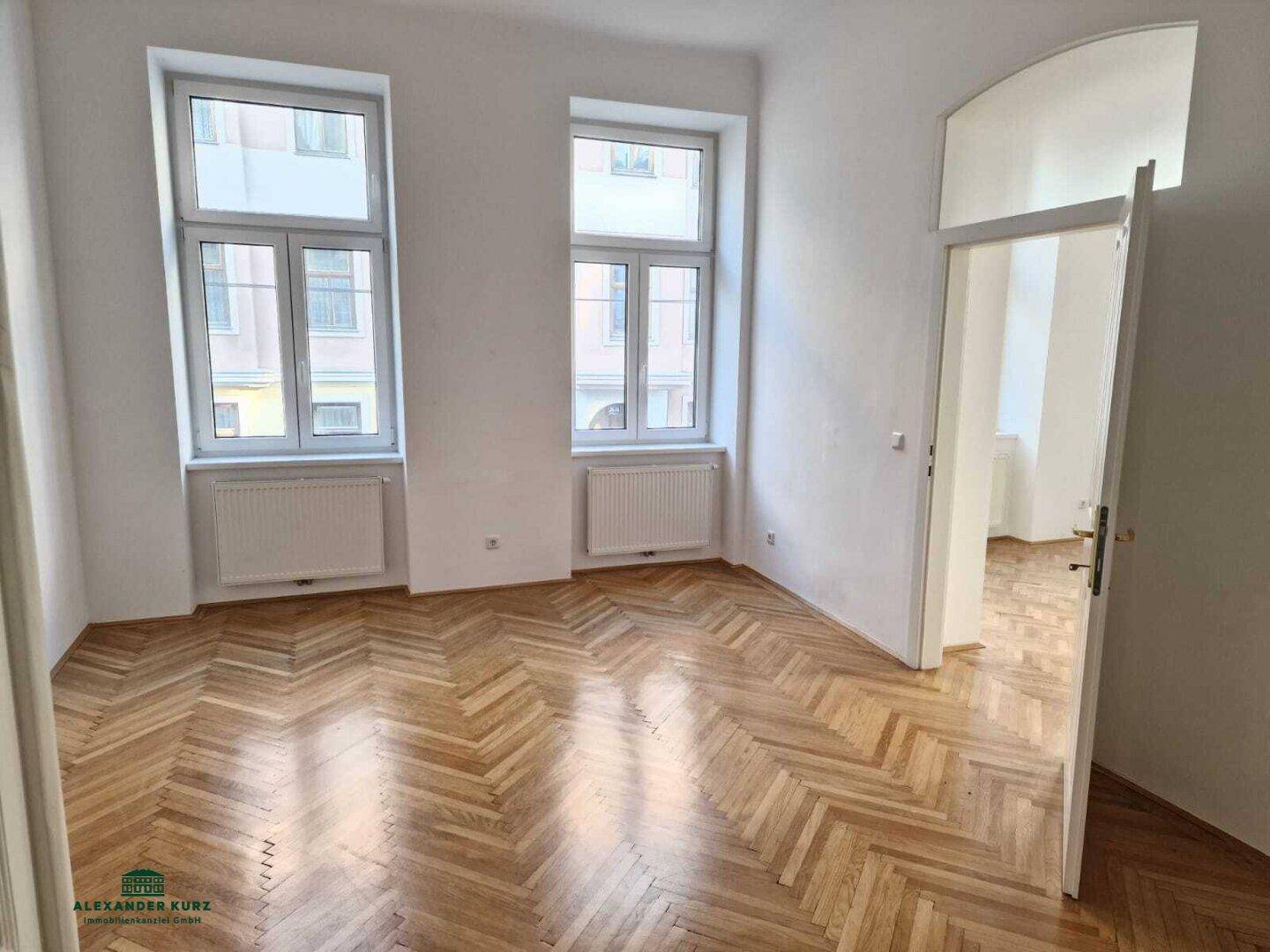 Leeres Zimmer mit Fischgrätparkett, hohen Fenstern und Heizkörpern unter den Fenstern.