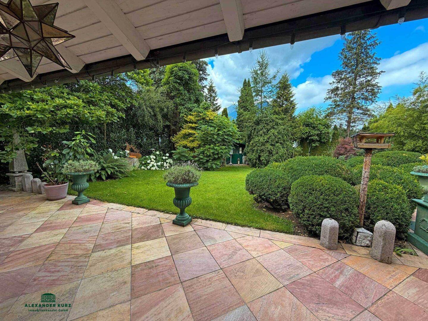 Geräumige Terrasse mit farbigen Steinplatten, umgeben von einem üppigen Garten und Blick auf die Berge.