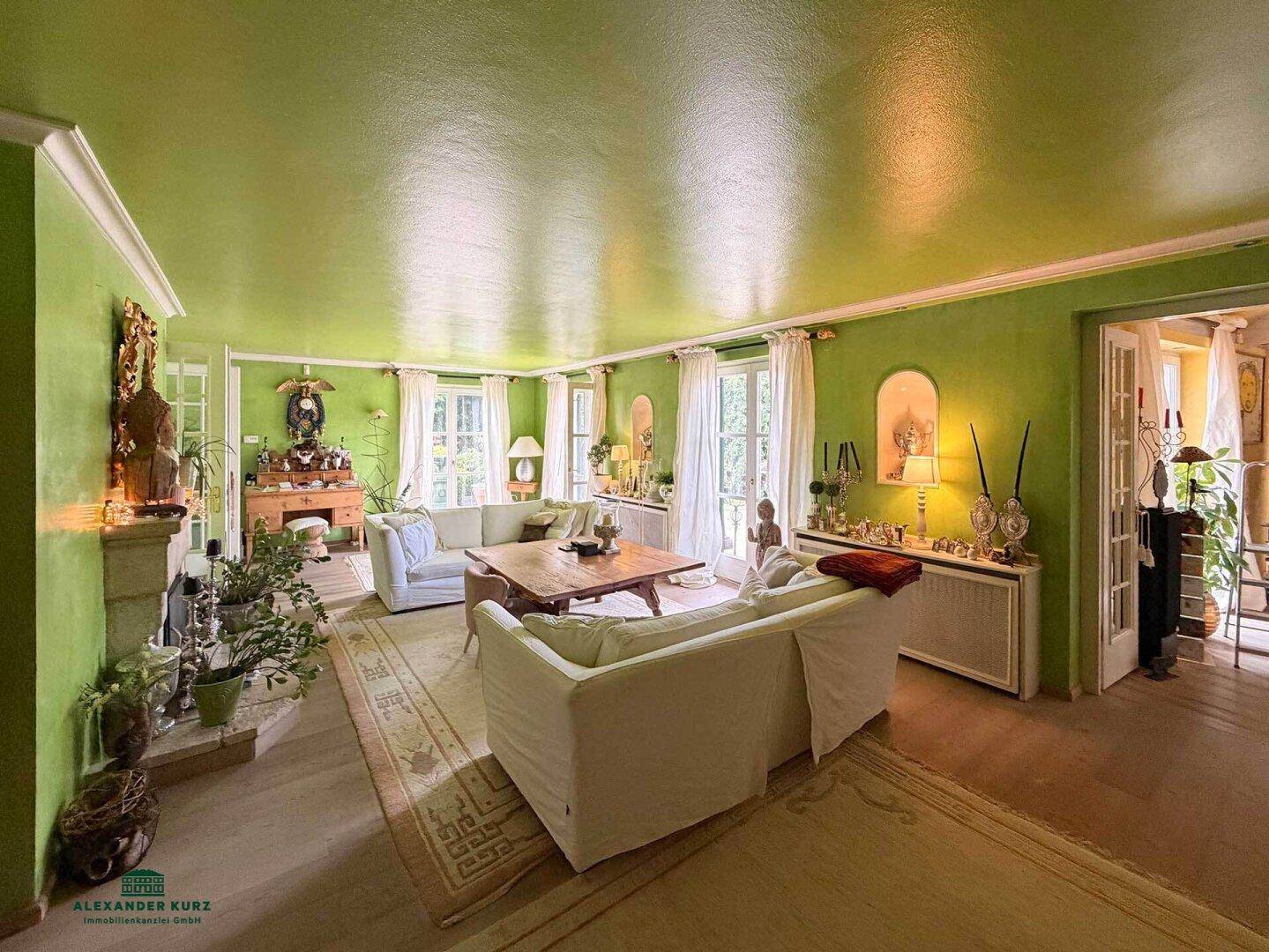 Gemütliches Wohnzimmer mit grünen Wänden, hellen Sofas, Holztisch und vielen Fenstern für natürliches Licht.