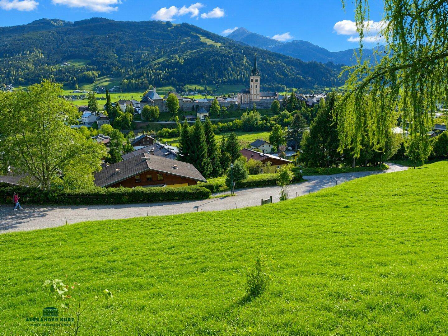 Panoramablick auf ein malerisches Dorf mit Kirche, umgeben von grünen Hügeln und Bergen.