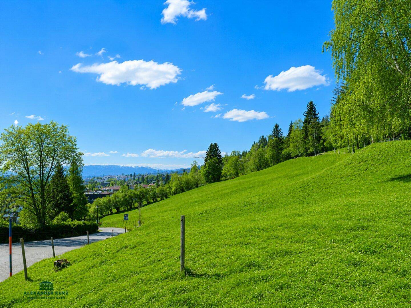 Sonnige grüne Wiese mit sanftem Hang und weitem Blick über eine Siedlung in der Ferne.