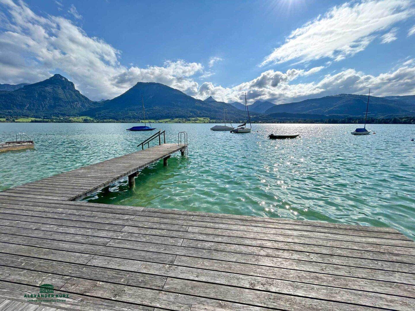 Langer Holzsteg führt in den See, umgeben von majestätischen Bergen und Segelbooten unter blauem Himmel.