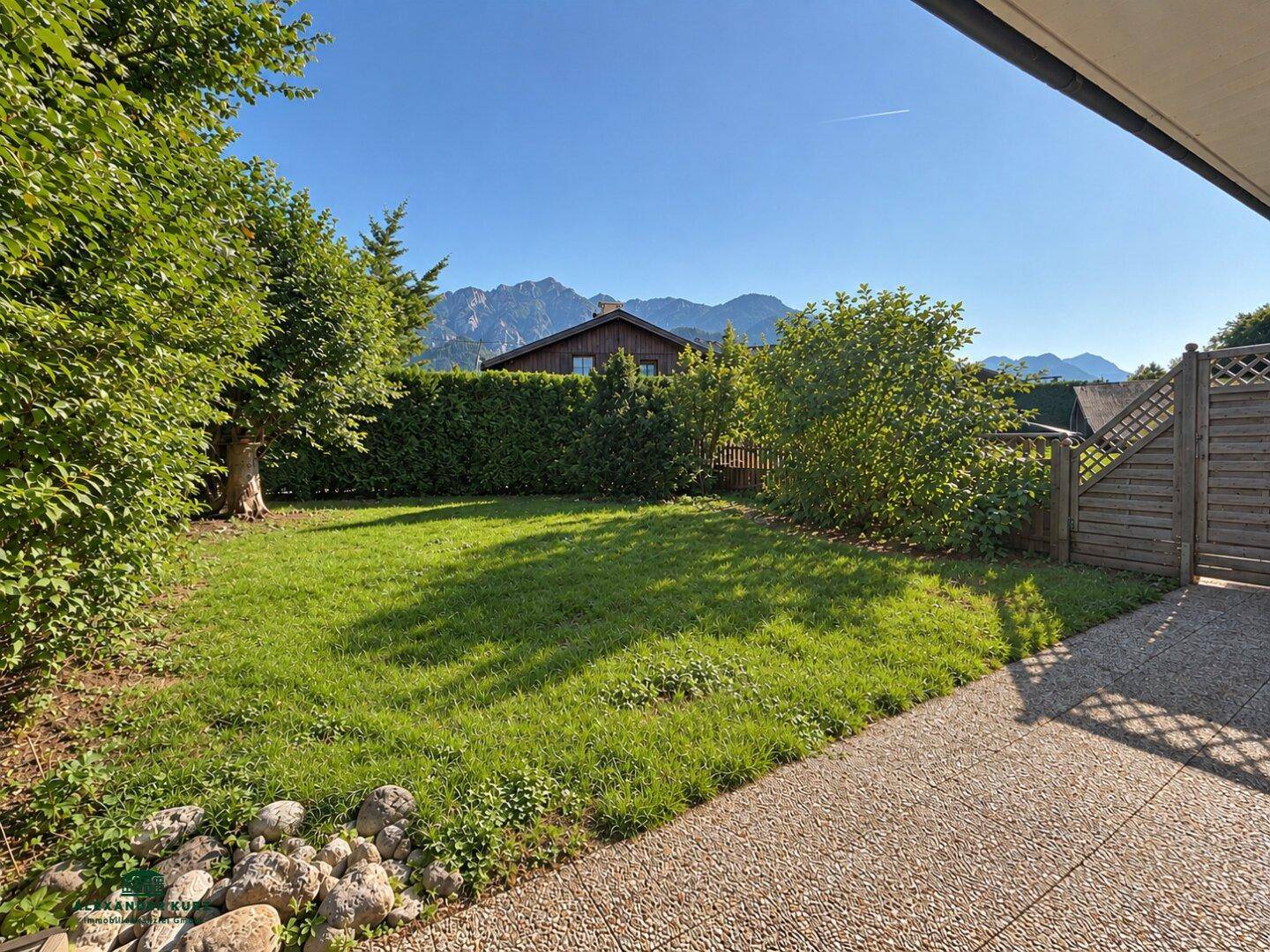 Gepflegter Garten mit Rasenfläche, Hecke und Bergblick im Hintergrund unter blauem Himmel.