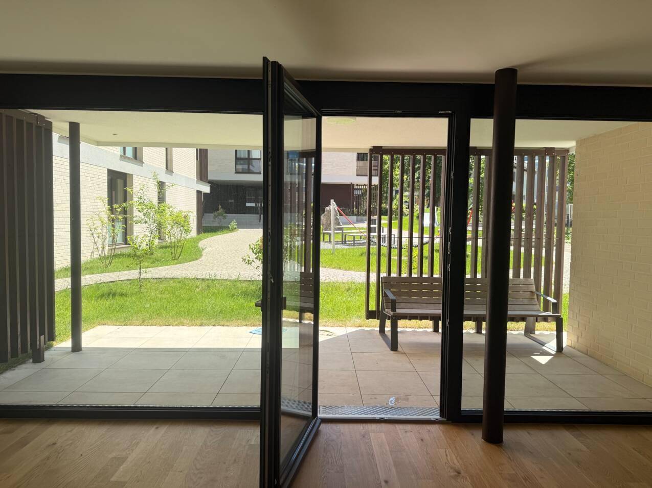 Heller Innenraum mit Holzboden und großen Glasfronten, die zu einer Terrasse und einem Gemeinschaftsgarten führen.