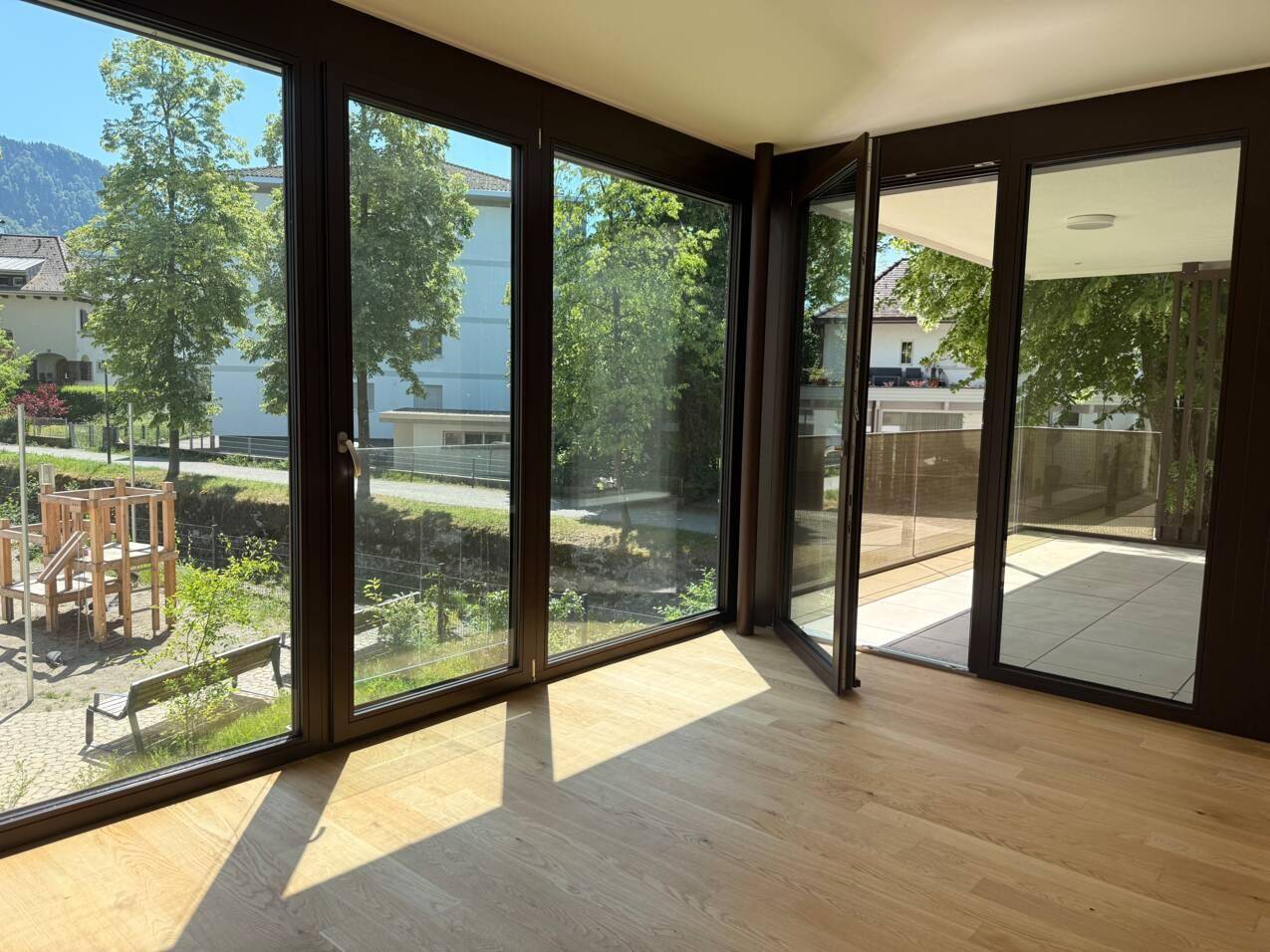 Lichtdurchfluteter Wohnraum mit großen Fenstern, Holzboden und Zugang zu einem sonnigen Balkon mit Grünblick.