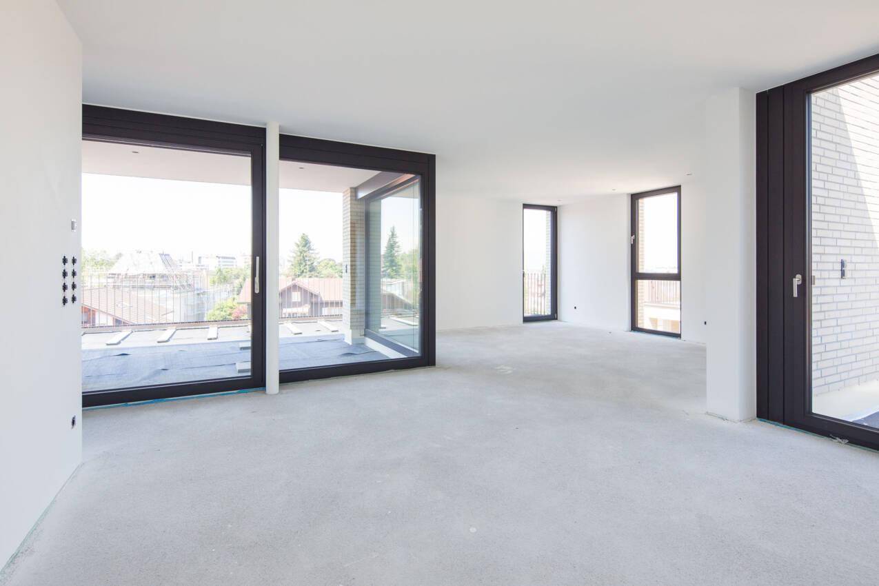 Geräumiger, unfertiger Wohnraum mit großen Fensterfronten und direktem Zugang zur Terrasse.