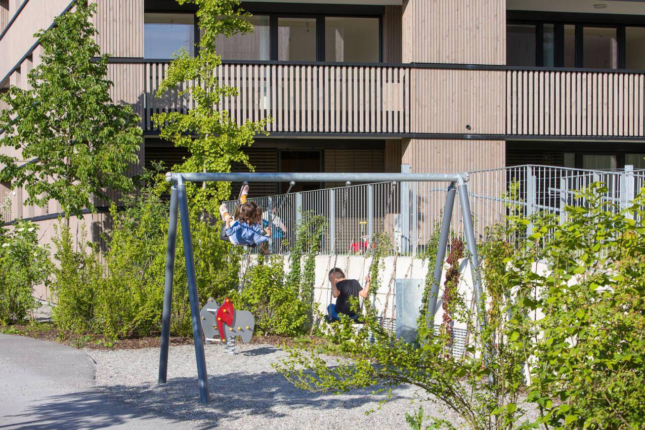 Kinderspielplatz mit Schaukeln und spielenden Kindern vor einer modernen Wohnanlage.