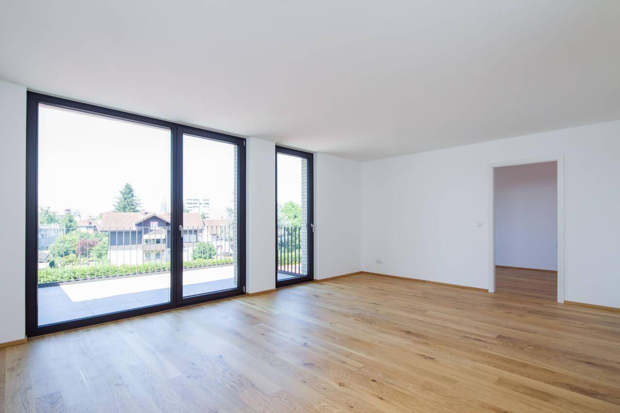Geräumiges Wohnzimmer mit Holzboden und großen Fenstern, die Zugang zu einem Balkon bieten.