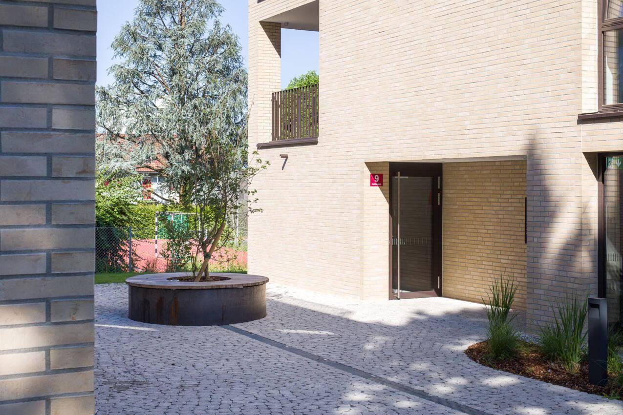 Eingangsbereich eines modernen Wohngebäudes mit gepflastertem Vorplatz und Baum.