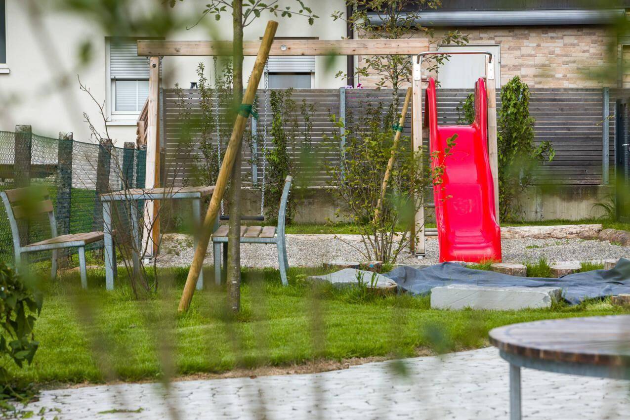 Kinderspielplatz im Gartenbereich mit roter Rutsche und Sitzgelegenheiten, umgeben von Grünflächen.
