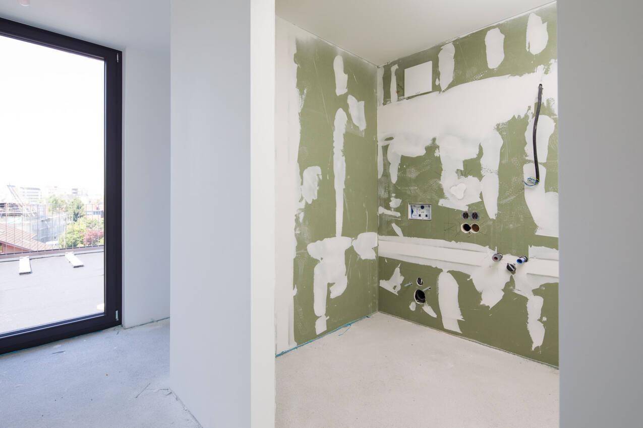 Unfertiges Badezimmer mit vorbereiteten Wänden, sichtbaren Installationen und einem Fenster.