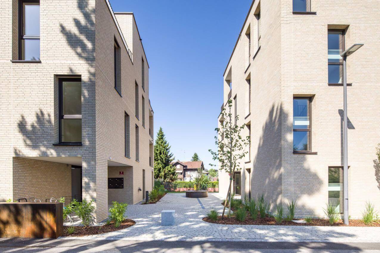 Blick zwischen zwei modernen Wohngebäuden mit gepflastertem Innenhof und Grünflächen.