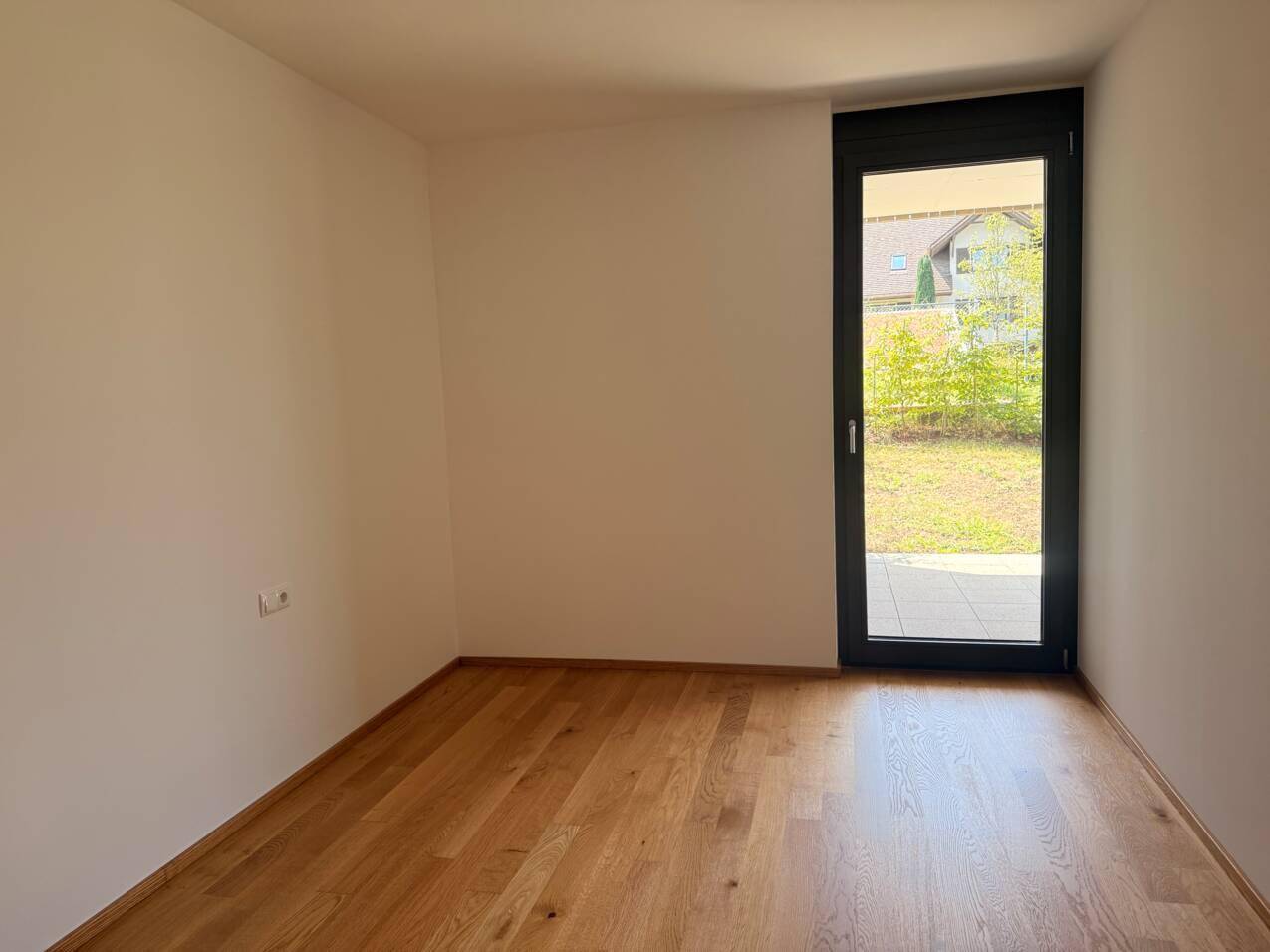 Leeres Zimmer mit Holzboden und Zugang zu einer Terrasse mit Blick ins Grüne.