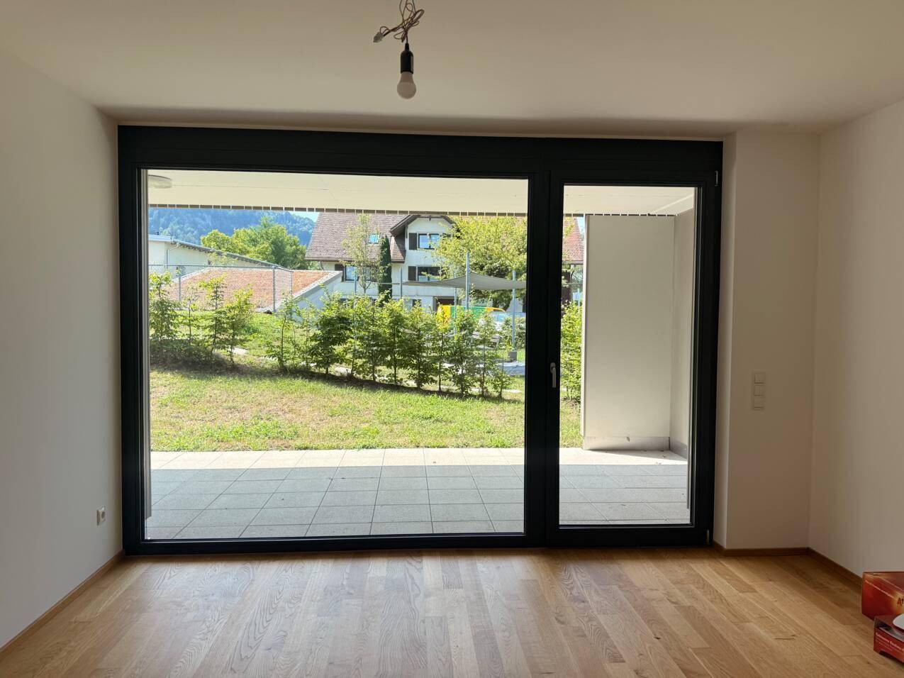 Heller Raum mit Holzboden und großer Schiebetür, die zu einer Terrasse und Grünfläche führt.