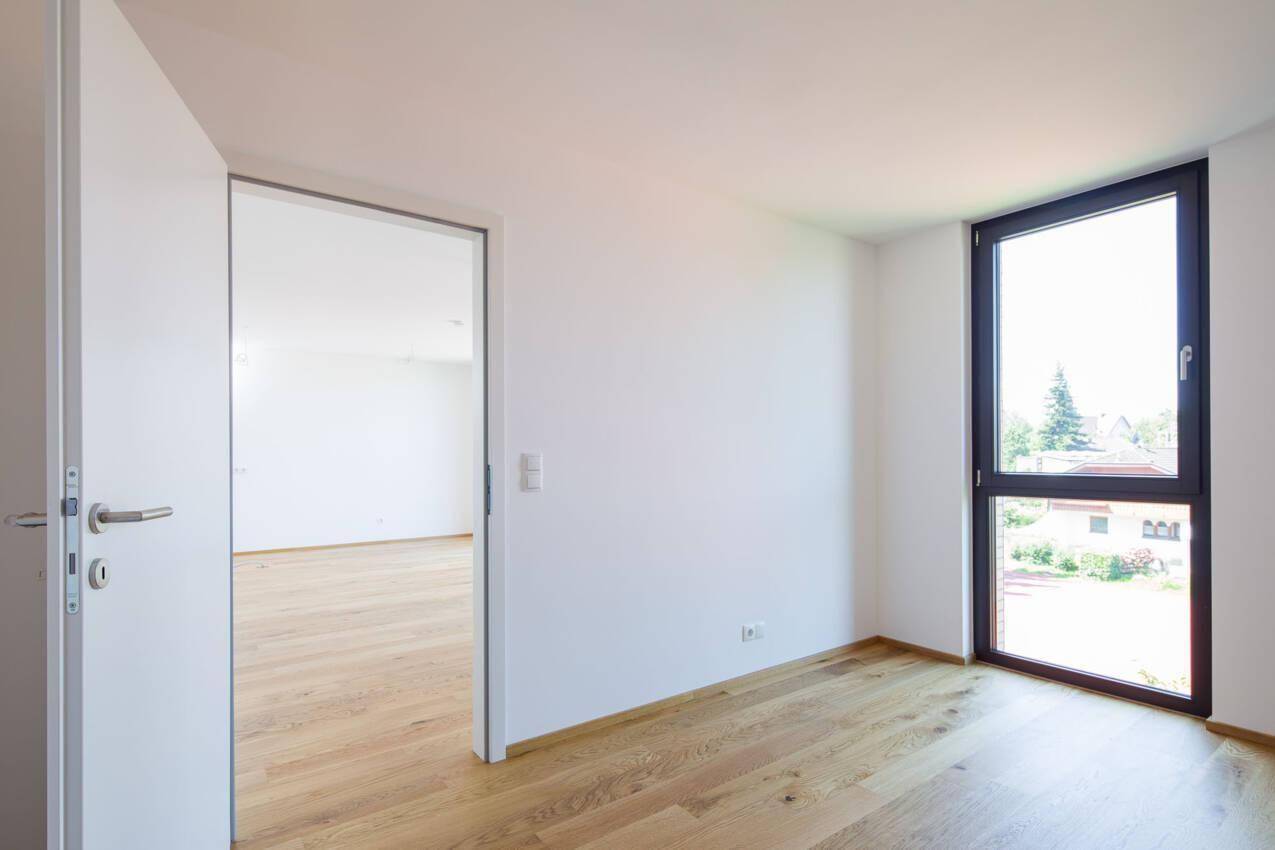 Leeres Zimmer mit Holzboden und großem Fenster, das viel Tageslicht hereinlässt.