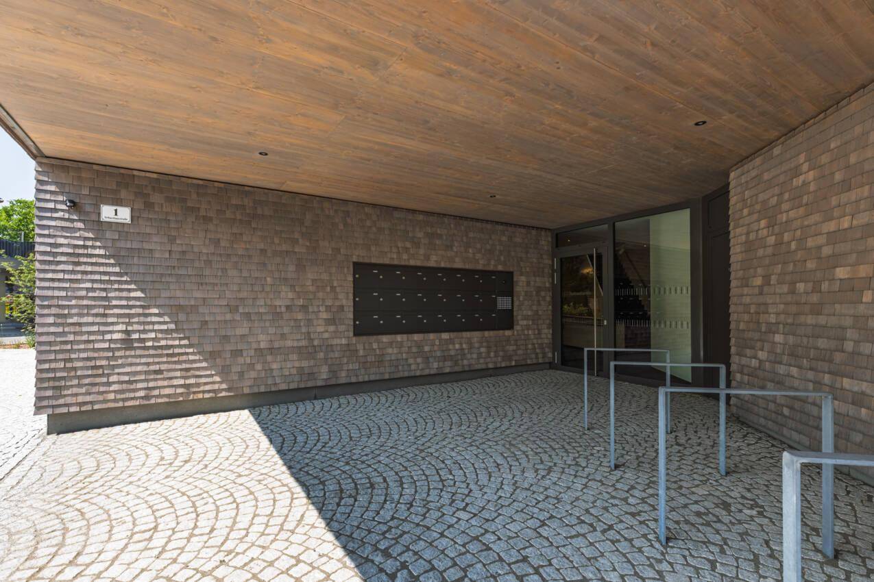 Eingangsbereich eines modernen Gebäudes mit Holzschindelfassade und gepflastertem Vorplatz.