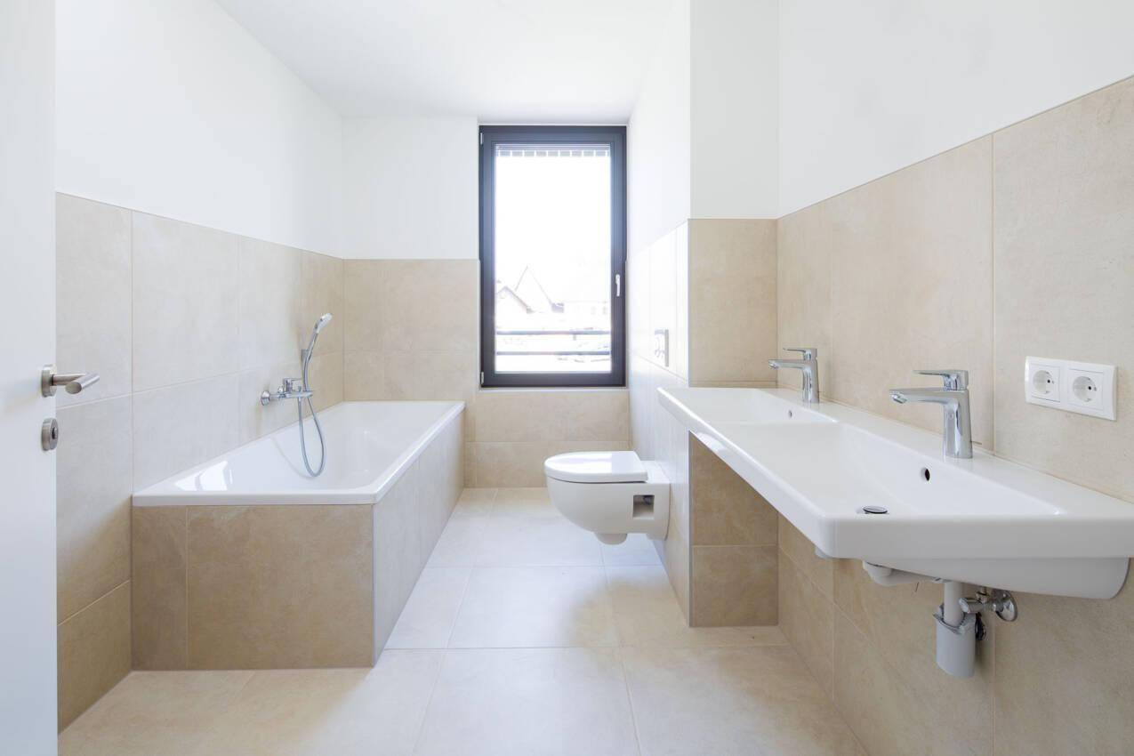 Modernes Badezimmer mit beigefarbenen Fliesen, Badewanne, wandhängendem WC und Doppelwaschbecken.