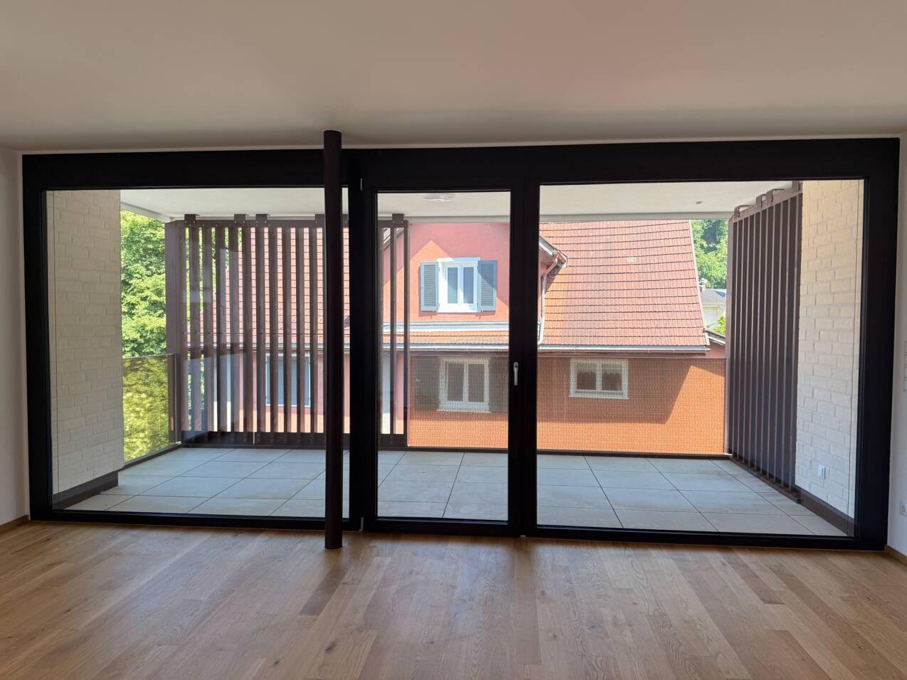 Heller Wohnraum mit Holzboden, großen Fenstern und Zugang zu einem Balkon mit Sichtschutz.