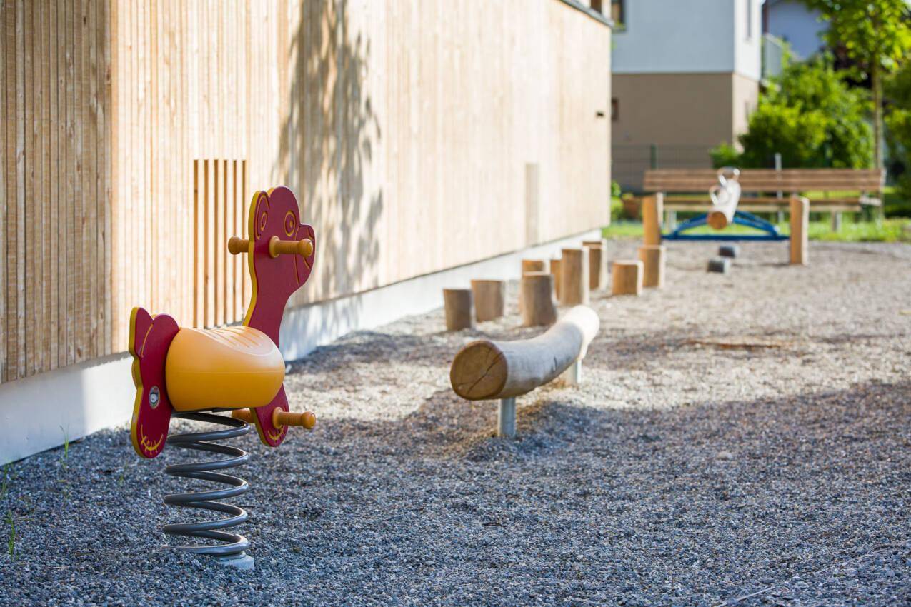 Kinderspielplatz mit Federwippe und Balancierbalken neben einem modernen Gebäude.