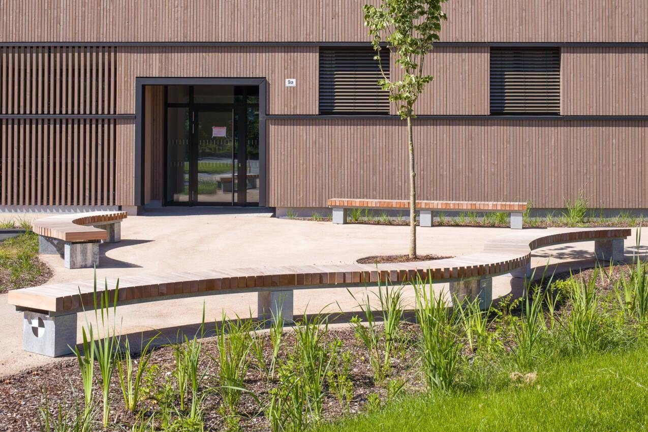 Eingangsbereich eines modernen Gebäudes mit Holzfassade und geschwungenen Sitzbänken im Freien.