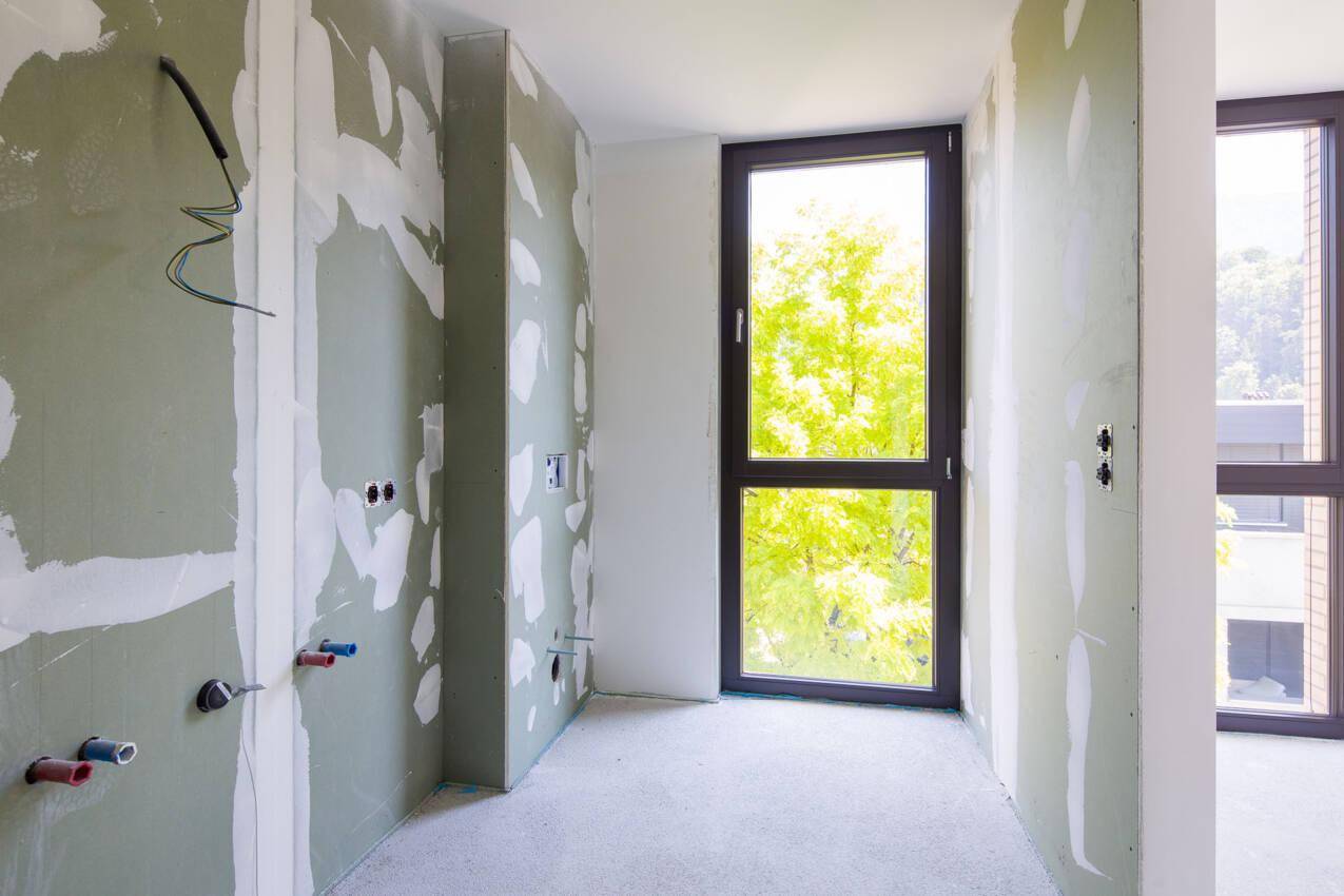 Unfertiges Badezimmer mit vorbereiteten Wänden, sichtbaren Installationen und Fenster mit Grünblick.