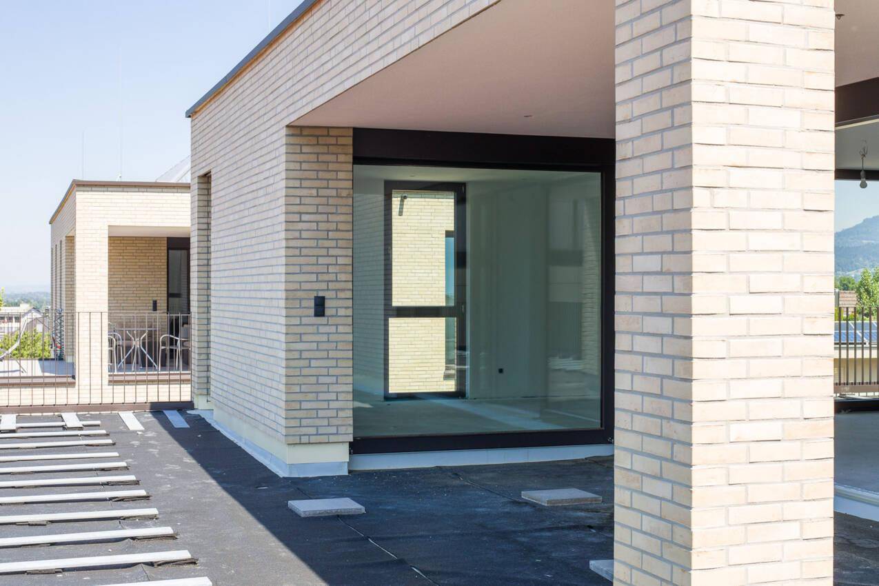 Dachterrasse im Bau mit heller Ziegelfassade und großen Fensterfronten, bietet weite Ausblicke.