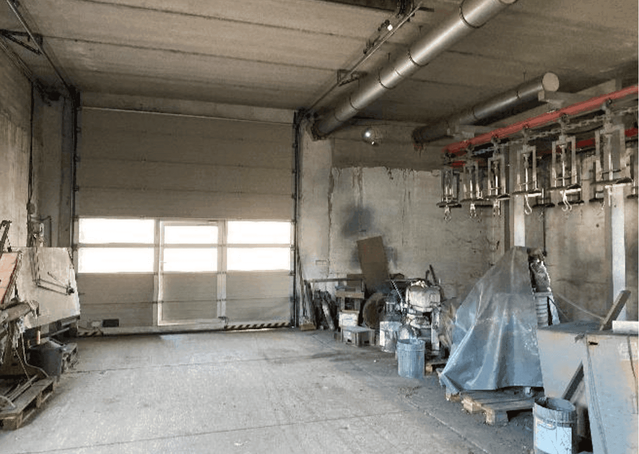 Geräumiger Lagerraum mit großem Rolltor, Betonboden und sichtbaren Lüftungsrohren an der Decke.