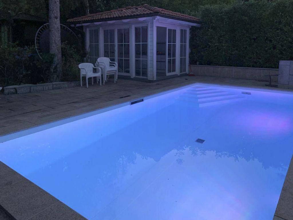 Beleuchteter Swimmingpool im Garten bei Nacht mit angrenzendem Gartenhaus und Sitzbereich.