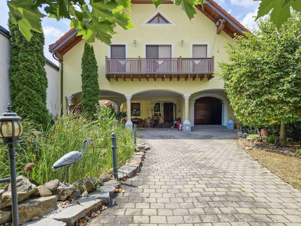 Vorderansicht des Hauses mit gepflasterter Auffahrt, kleinem Teich und Balkon im Obergeschoss.