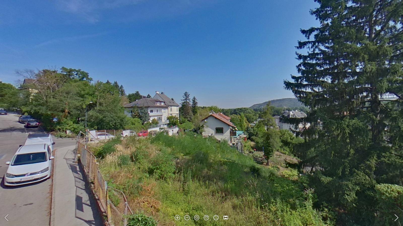 Blick auf eine Wohnstraße mit parkenden Autos, umgeben von grünen Hügeln und Häusern.