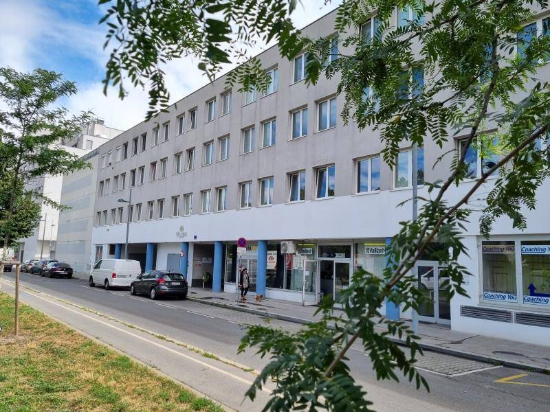 Moderne Gebäudefassade mit vielen Fenstern und Geschäften im Erdgeschoss, umgeben von Bäumen.