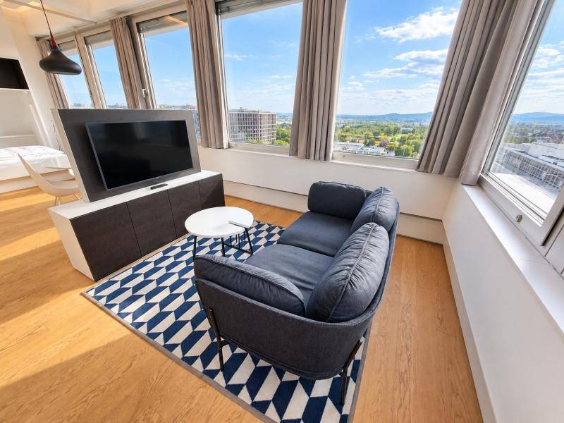Helles Wohnzimmer mit bequemer Couch, Fernseher und weitem Blick über die Stadt und die grüne Landschaft.
