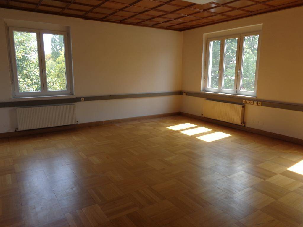 Heller Raum mit Fischgrätparkett, Holzdecke und zwei großen Fenstern, die viel Tageslicht hereinlassen.