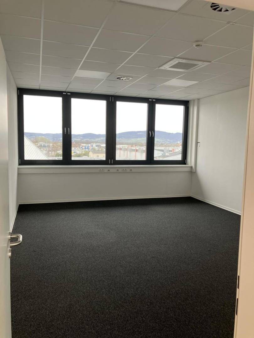 Helles Büro mit Teppichboden und großen Fenstern, die viel Tageslicht hereinlassen.