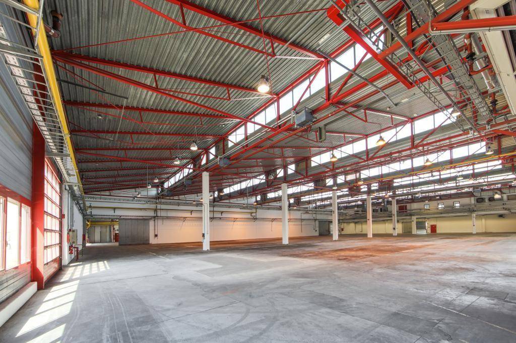 Weitläufige Lagerhalle mit roter Stahlkonstruktion, Oberlichtern und Betonboden.