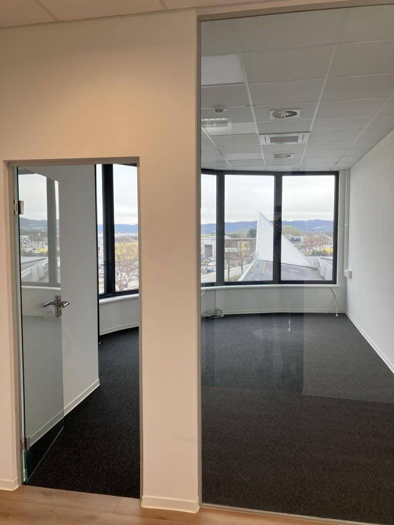 Leeres Büro mit Teppichboden, großen Fenstern und Blick auf die Umgebung.