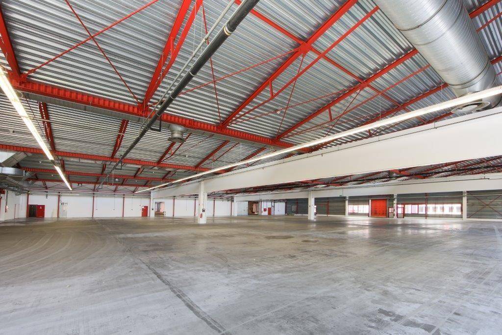 Geräumige Lagerhalle mit Betonboden, roter Stahlkonstruktion und Lüftungsrohren an der Decke.