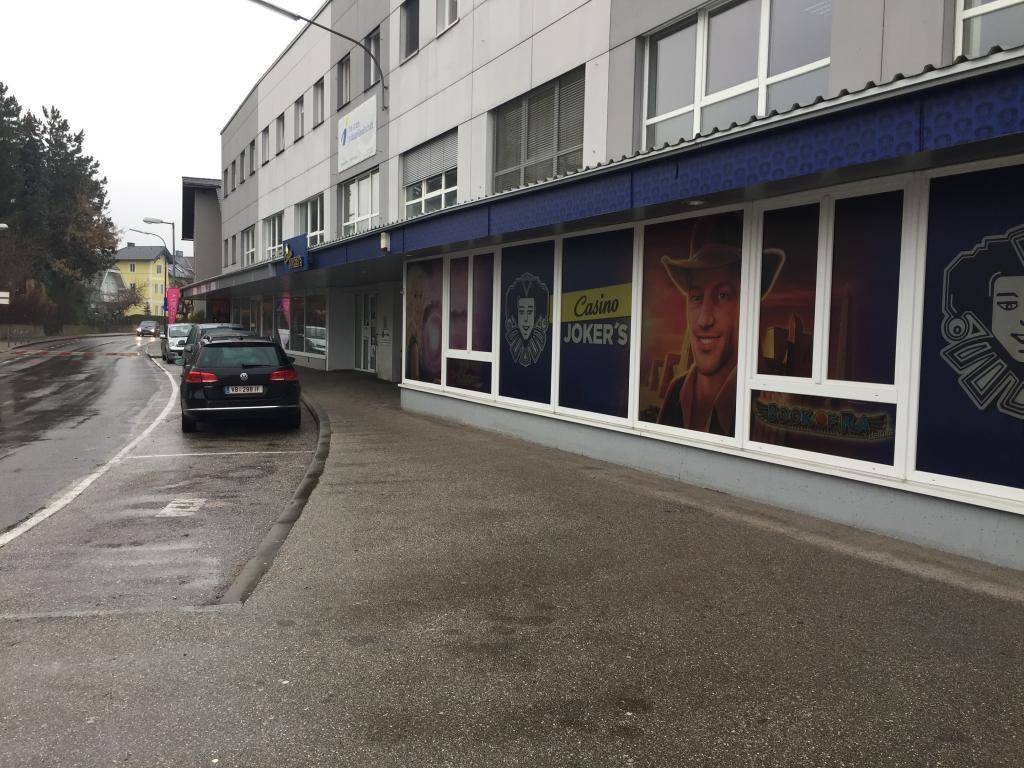 Straßenansicht des Geschäftsgebäudes mit großen Schaufenstern und Werbeplakaten an der Fassade, mit geparkten Autos.