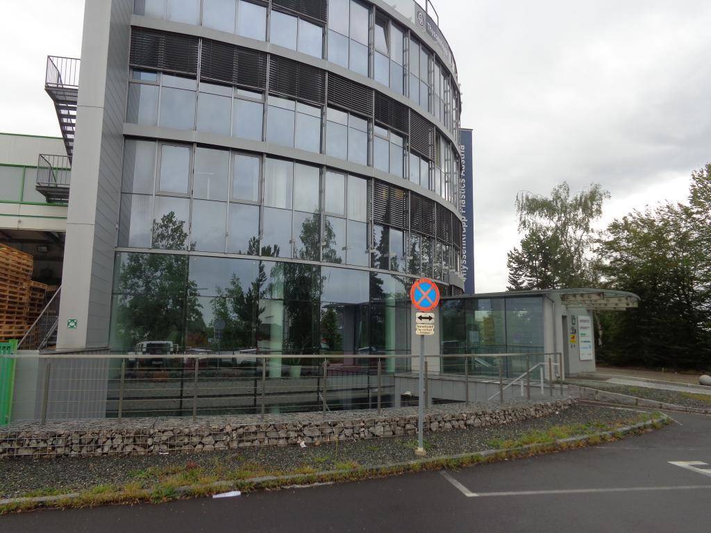Moderne Bürogebäude-Fassade mit geschwungenen Glasfronten und reflektierenden Fenstern, umgeben von Grünflächen und einem Parkplatz.