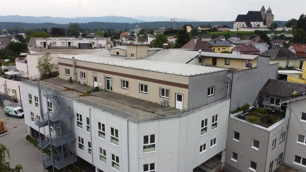 Luftaufnahme eines Gebäudekomplexes mit verschiedenen Dächern und einer Kirche im Hintergrund der Stadt, umgeben von Grünflächen.