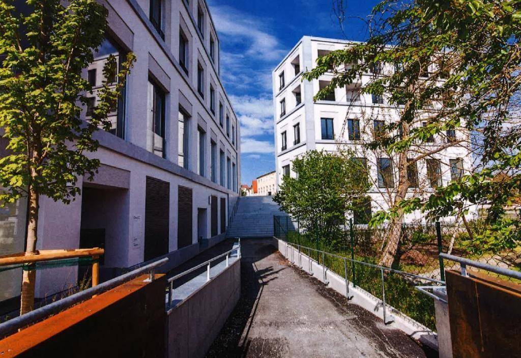 Eine moderne Rampe führt zwischen zwei weißen Gebäuden hindurch, umgeben von jungem Grün und Metallgeländern.