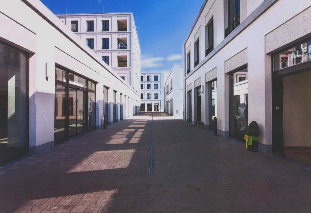 Eine moderne Passage zwischen weißen Gebäuden mit großen Schaufenstern und langen Schatten auf dem Boden.