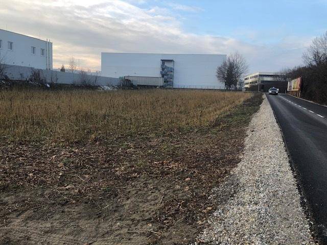 Blick über das unbebaute Grundstück mit trockener Vegetation und Industriegebäuden im Hintergrund.