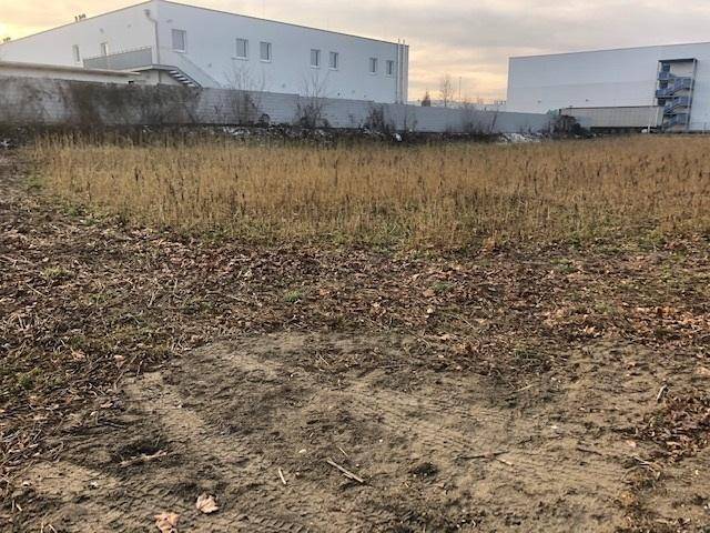 Unbebautes Grundstück mit trockener Erde und Gras, umgeben von Gewerbebauten unter bewölktem Himmel.