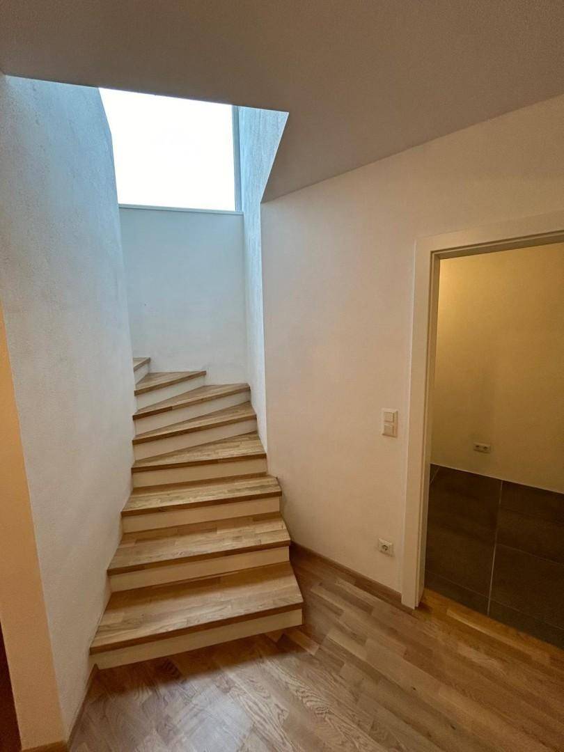 Treppenaufgang mit Holzstufen und weiß gestrichenen Wänden, hell durch Oberlicht.