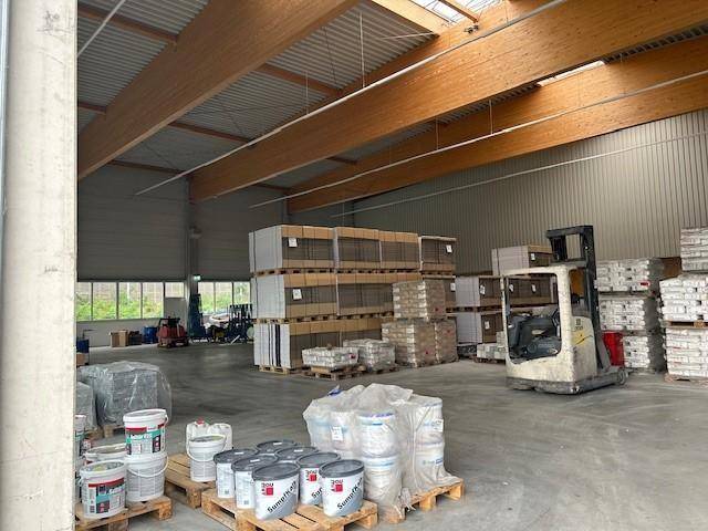 Lagerhalle mit gestapelten Waren, Farbbehältern im Vordergrund und einem Gabelstapler, mit Fenstern im Hintergrund.