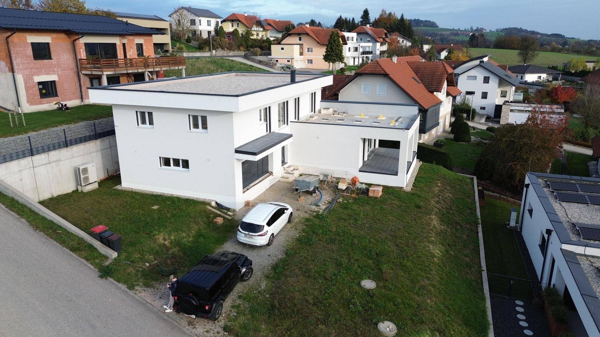 Modernes weißes Haus im Bau mit Flachdach und umliegender Landschaft unter bewölktem Himmel.