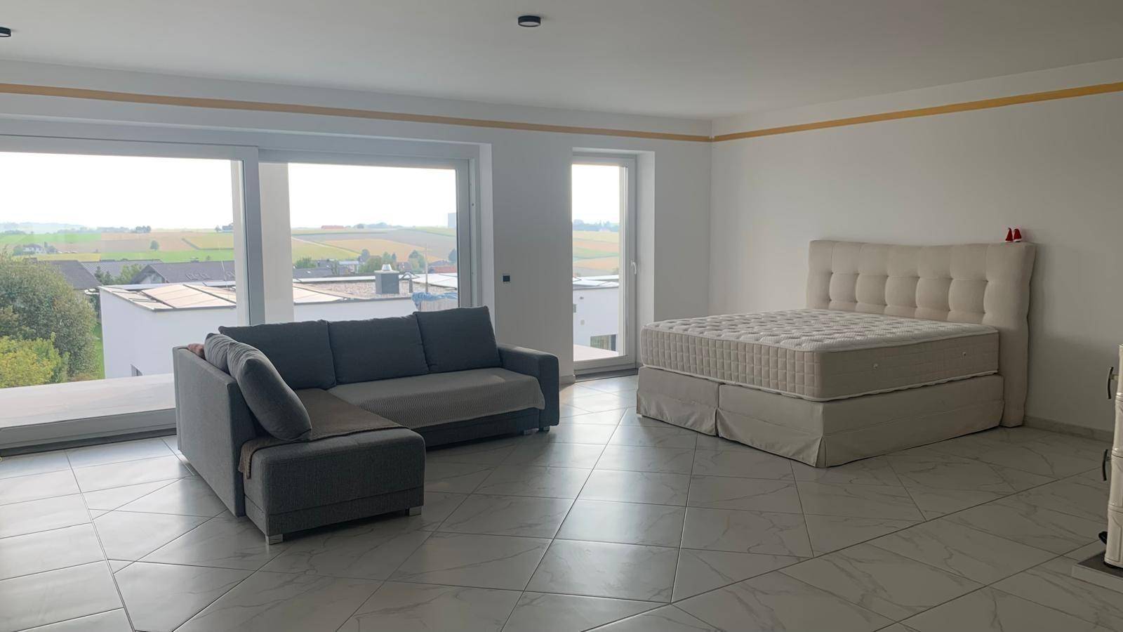 Heller Wohnbereich mit Sofa und Bett, großen Fenstern und weitem Blick ins Grüne.