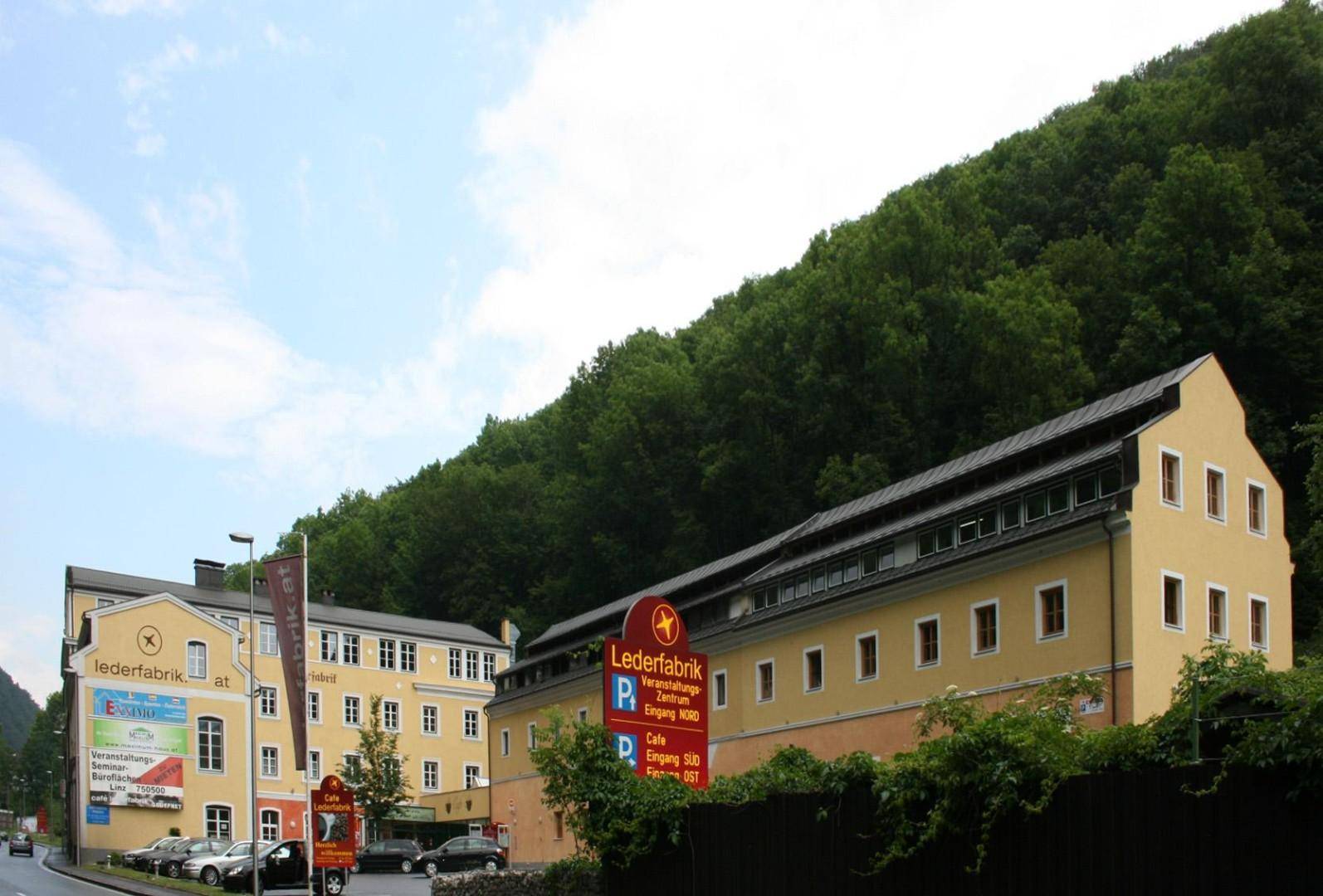 Historisches Gebäude mit gelber Fassade am Fuße eines bewaldeten Hügels, umgeben von Natur.