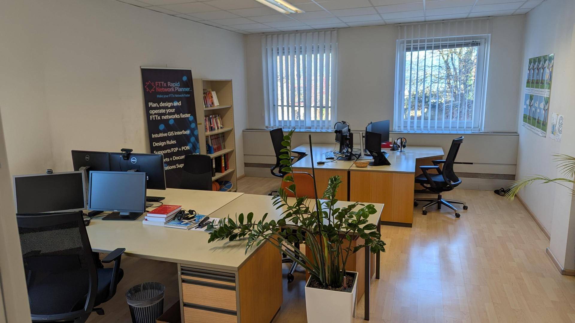 Großes Büro mit mehreren Arbeitsplätzen, Bücherregal und Pflanzen, gut beleuchtet durch Fenster.